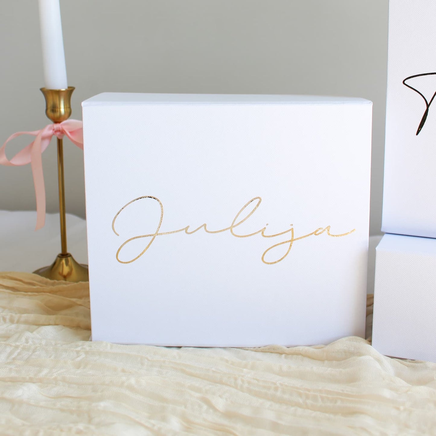 A white rectangular bridesmaid proposal gift box with the name 'Julija' written in elegant gold cursive script, styled on a beige textured fabric next to a gold candlestick holder with a pink ribbon.