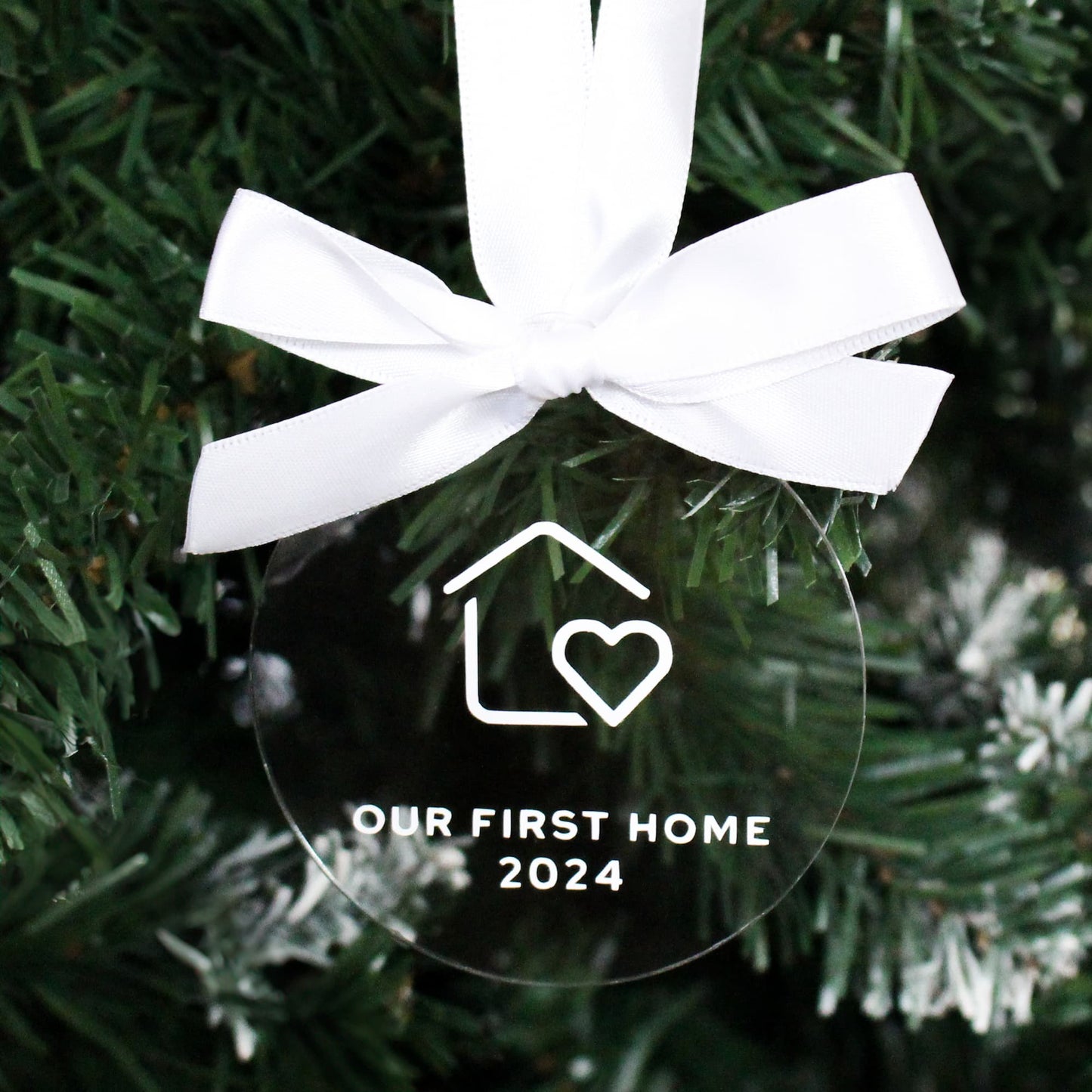Clear ornament with white ribbon and 'Our First Home 2024' text on a Christmas tree.