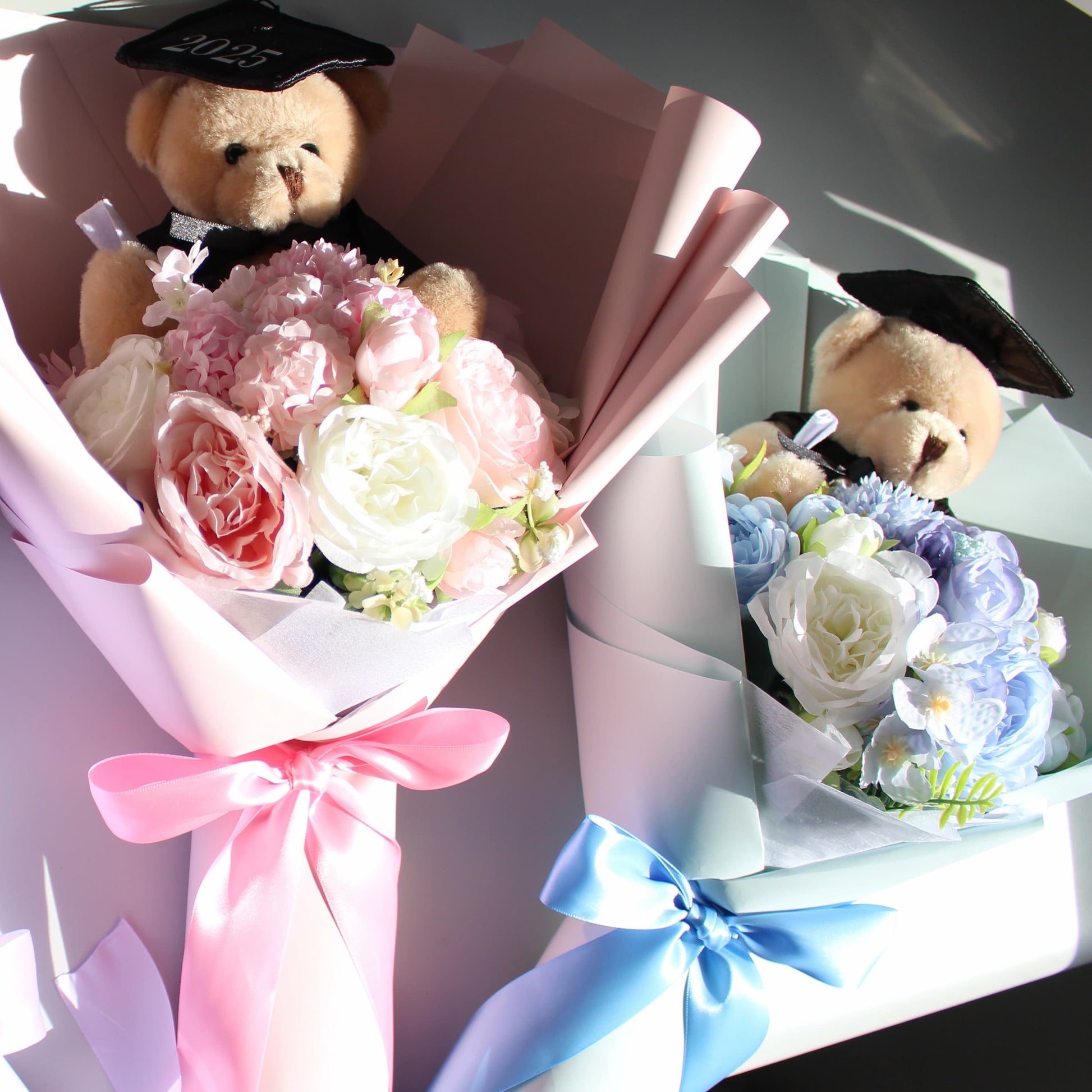 Two teddy bears in graduation caps with pink and blue flower bouquets