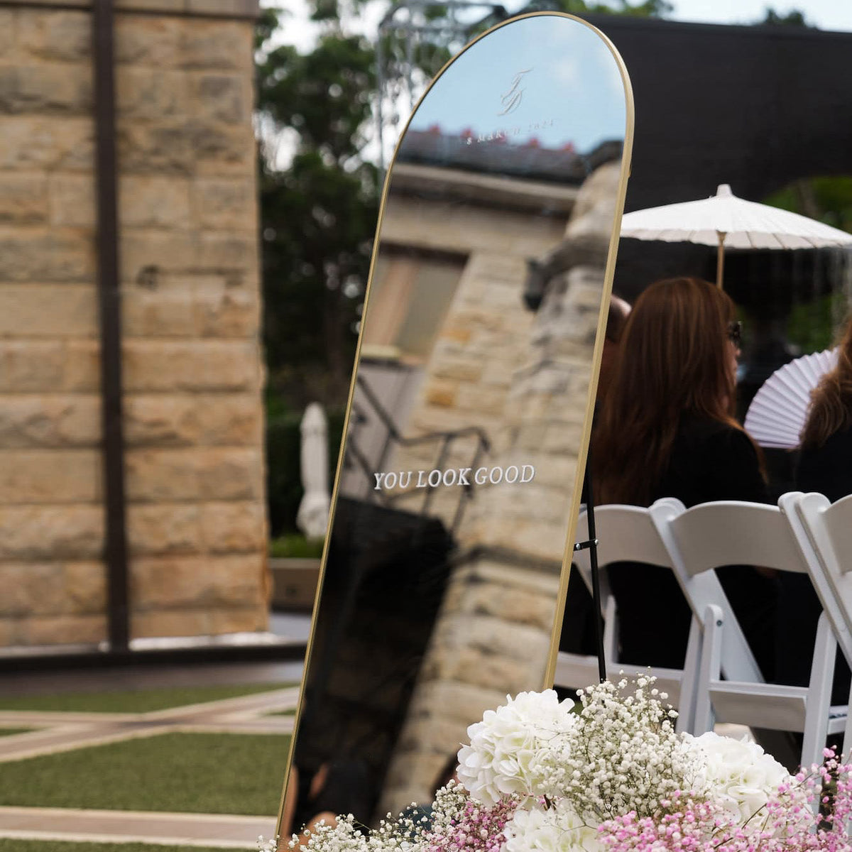 Decorative mirror with 'YOU LOOK GOOD' text in an outdoor setting with chairs and flowers.