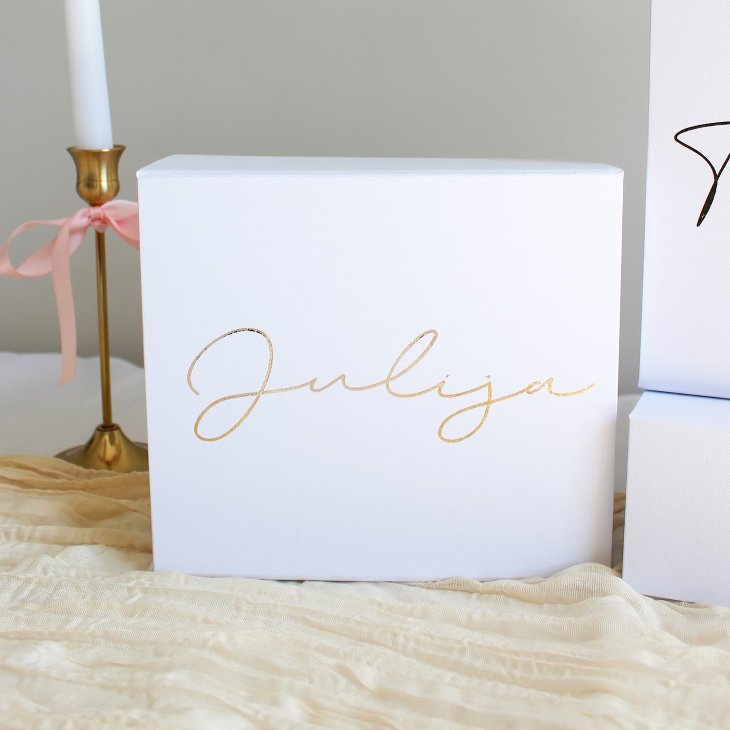 A white rectangular bridesmaid proposal gift box with the name 'Julija' written in elegant gold cursive script, styled on a beige textured fabric next to a gold candlestick holder with a pink ribbon.