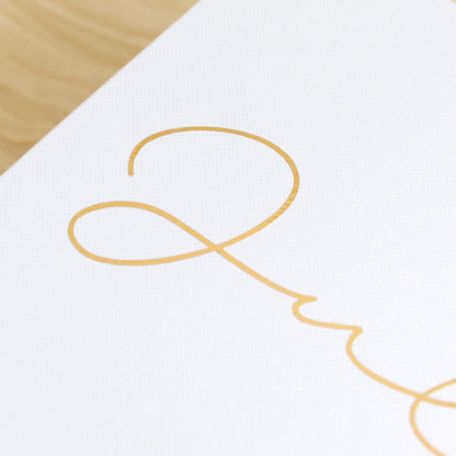 A close-up of a white textured bridesmaid gift box featuring a delicate gold cursive script, with a light wood surface visible in the top-left corner.
