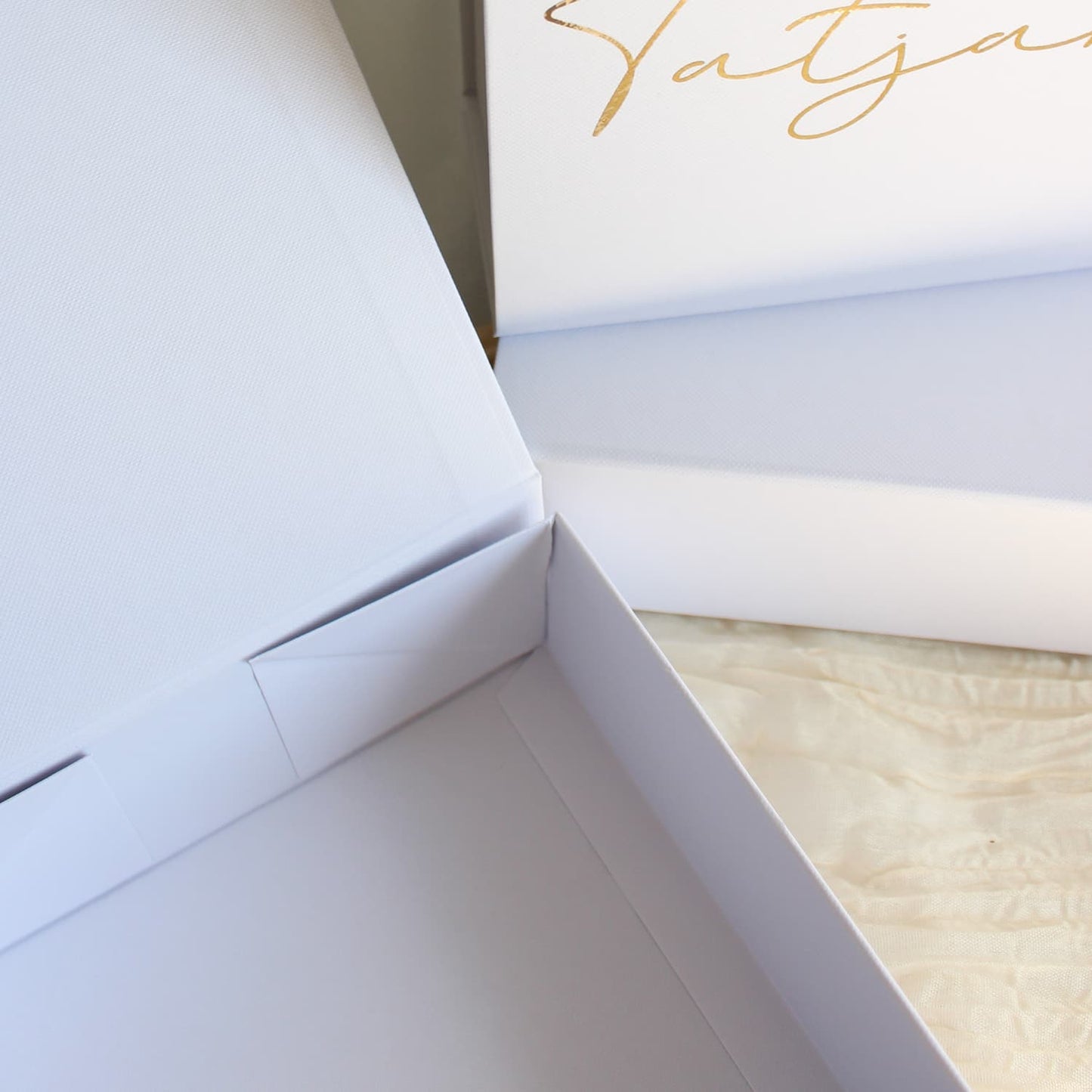 A close-up of a white gift box with a hinged lid, featuring elegant gold foil script lettering on the inside of the lid. The box is shown open, revealing a light beige textured fabric lining inside.