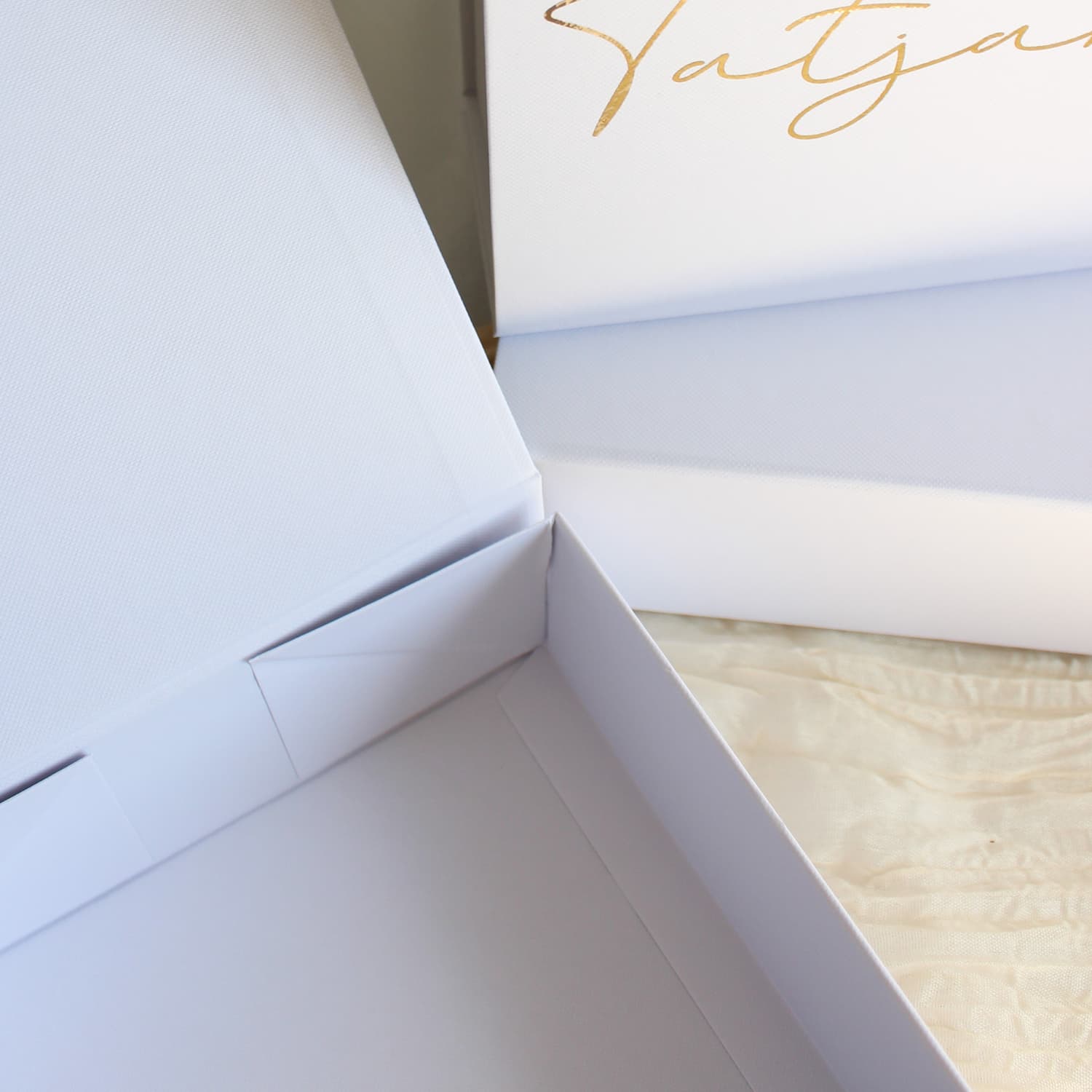 A close-up of a white gift box with a hinged lid, featuring elegant gold foil script lettering on the inside of the lid. The box is shown open, revealing a light beige textured fabric lining inside.