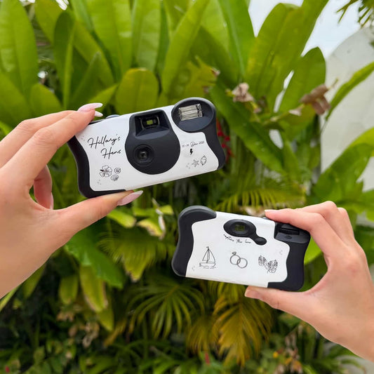 Two hands holding Hillary's Hen's instant cameras against a green leafy background