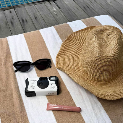 Sunglasses, disposable camera, hat, and lip balm on a striped towel by a poolside.