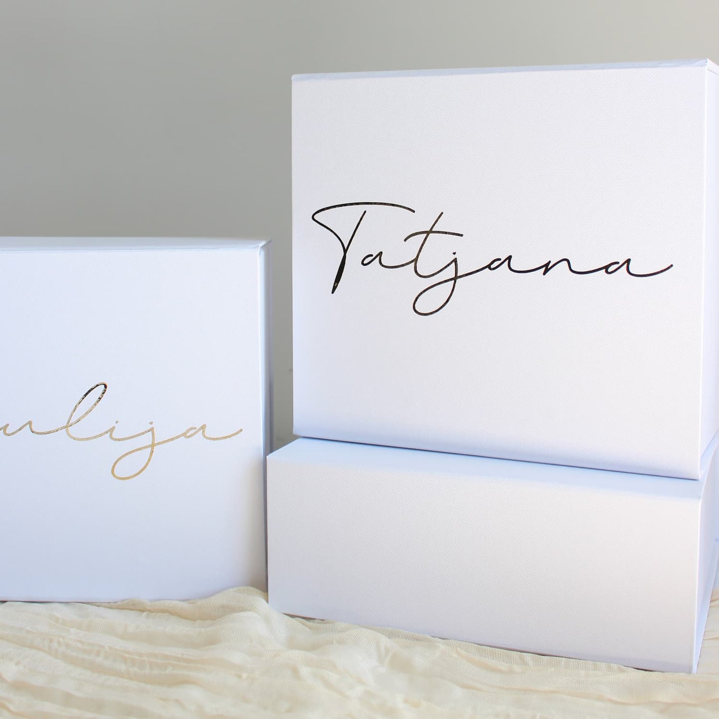 Three white rectangular gift boxes with personalized script names printed on the front, displayed on a cream textured fabric against a neutral gray background.