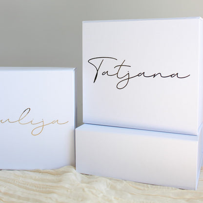 Three white rectangular gift boxes with personalized script names printed on the front, displayed on a cream textured fabric against a neutral gray background.