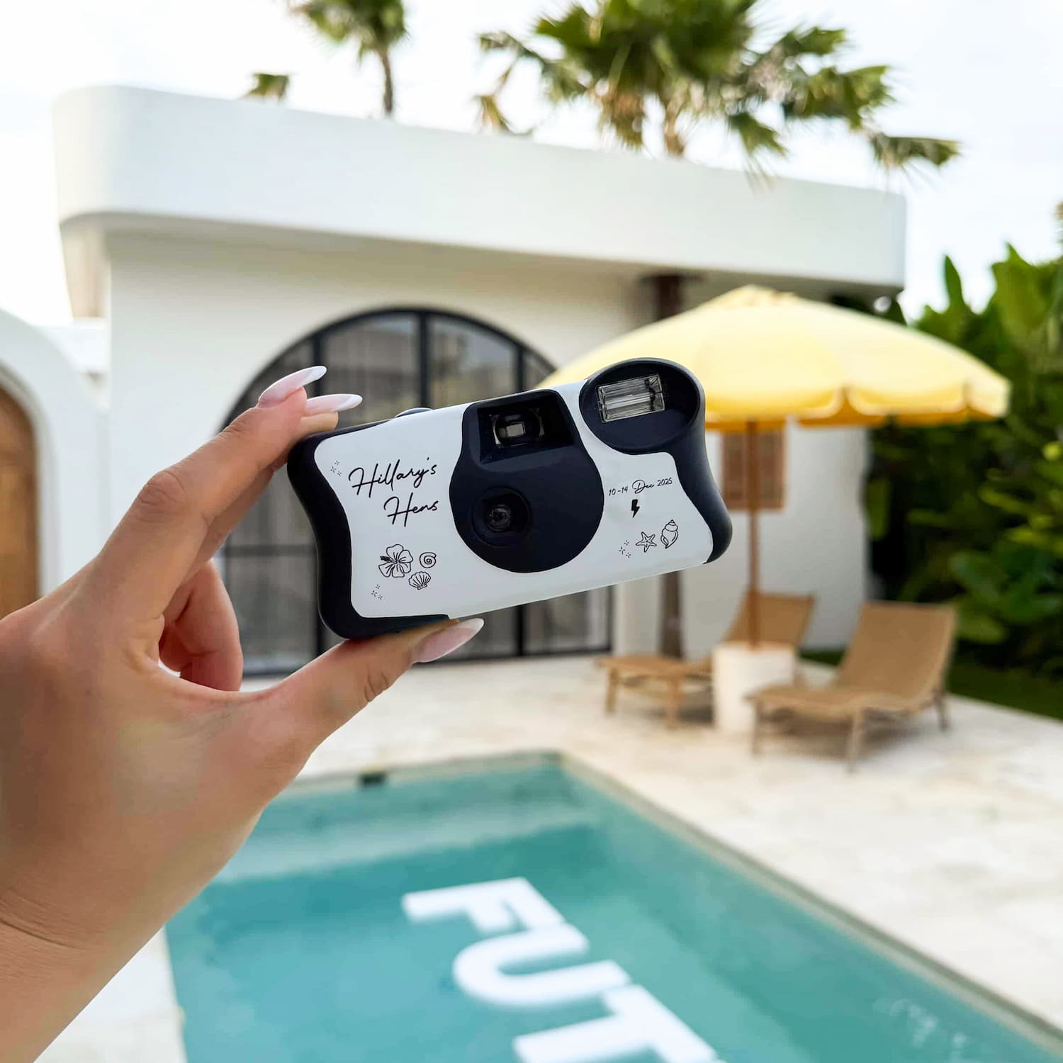 Hand holding a disposable camera with a pool and lounge chairs in the background