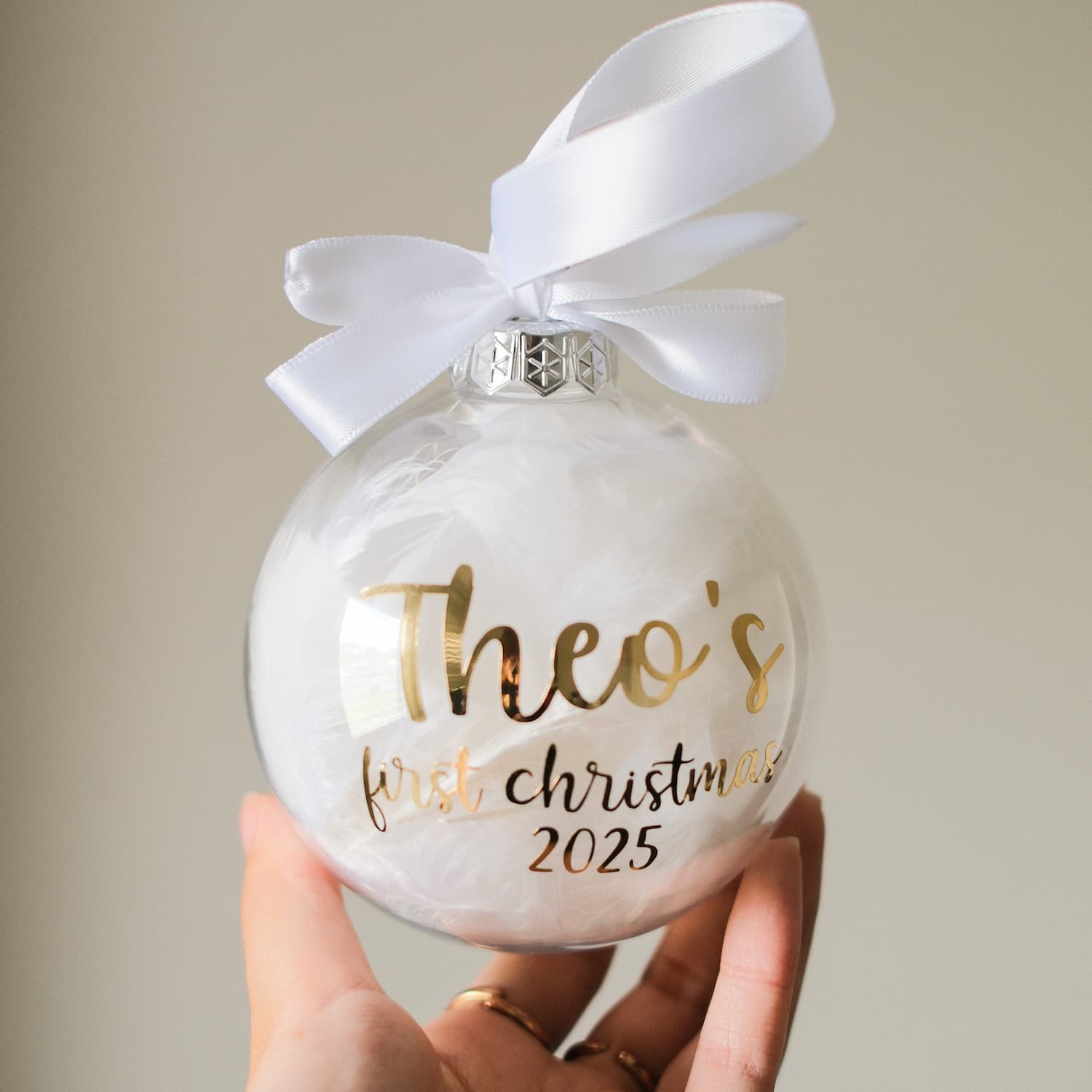 Feather Christmas ornament with gold text 'Theo's first Christmas 2025' and a white ribbon, held by a hand against a neutral background.