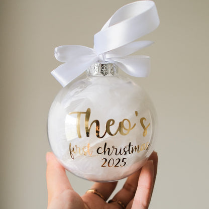 Feather Christmas ornament with gold text 'Theo's first Christmas 2025' and a white ribbon, held by a hand against a neutral background.
