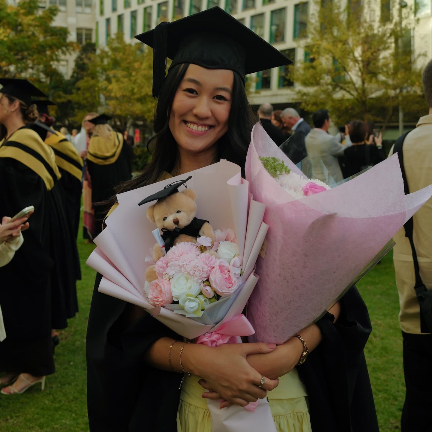 Graduate holding a bouquet of flowers and a teddy bear plush toy in a celebratory setting.