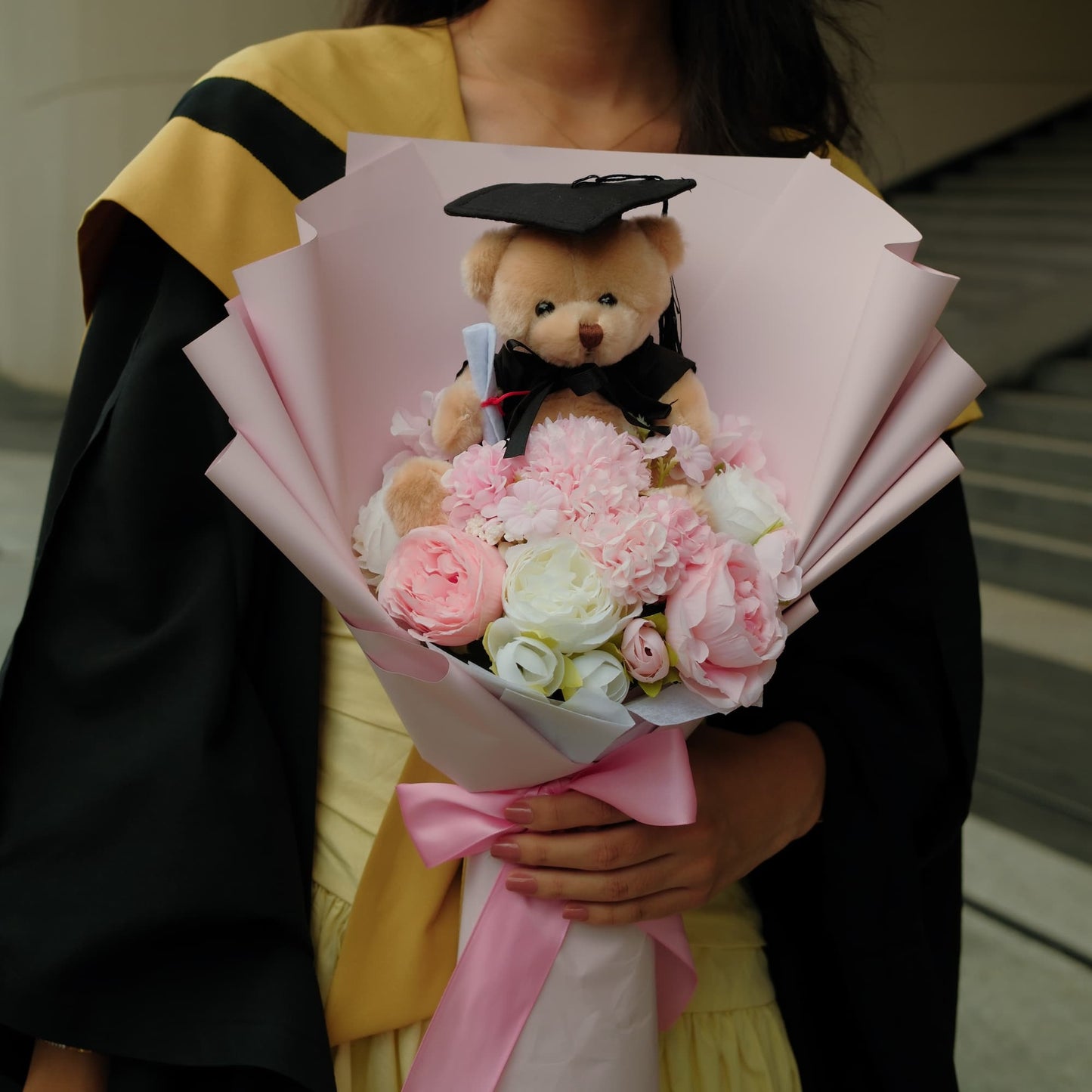 Person holding a bouquet of flowers with a teddy bear wearing a graduation cap.