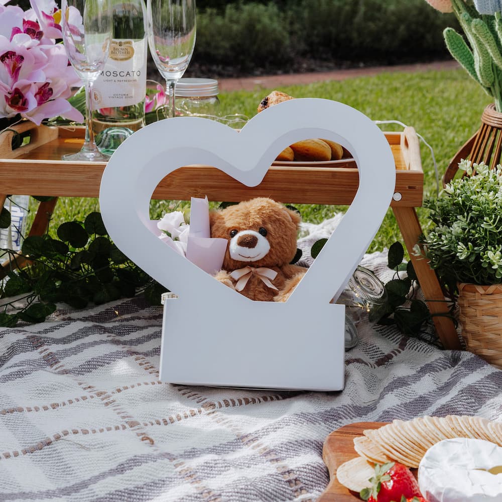White heart-shaped box with a teddy bear on a picnic blanket with wine and snacks.