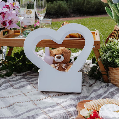 White heart-shaped box with a teddy bear on a picnic blanket with wine and snacks.