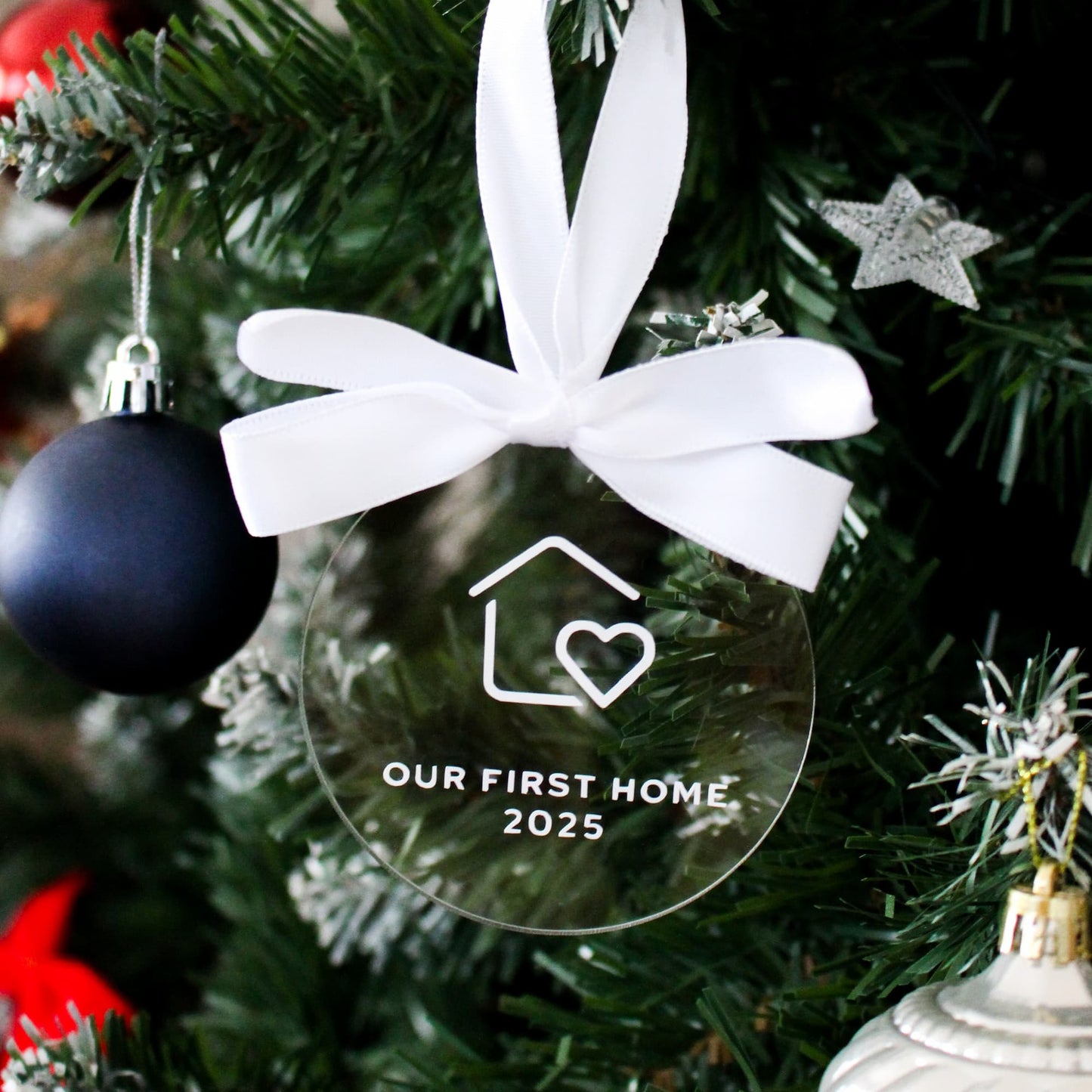 Clear ornament with white ribbon and 'Our First Home 2025' text on a Christmas tree.