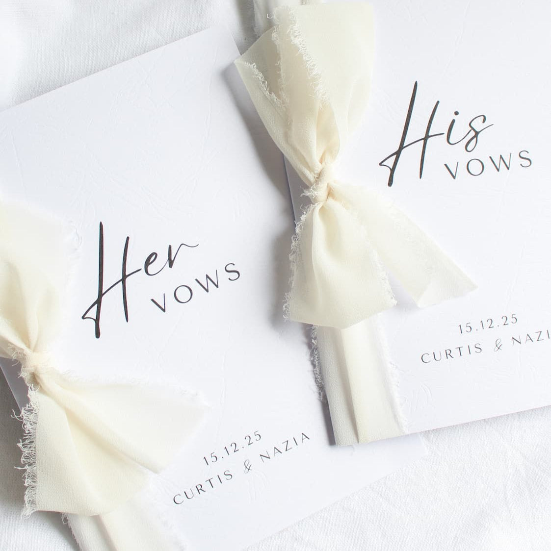 Two vow books titled 'Her Vows' and 'His Vows' with ribbons on a white fabric background.