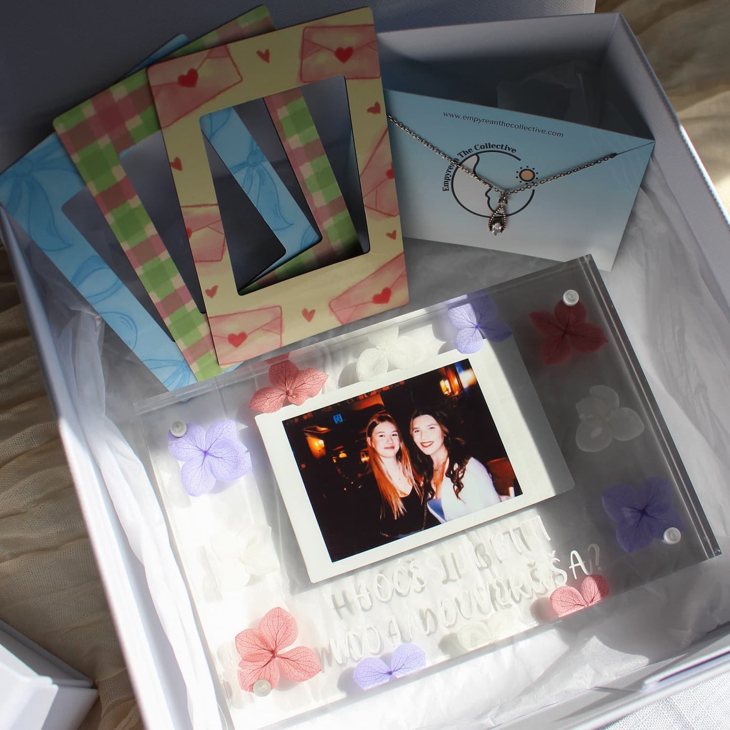 A gift box containing a personalized acrylic photo frame with preserved flowers, a blue necklace, and colorful envelope-shaped picture frames.
