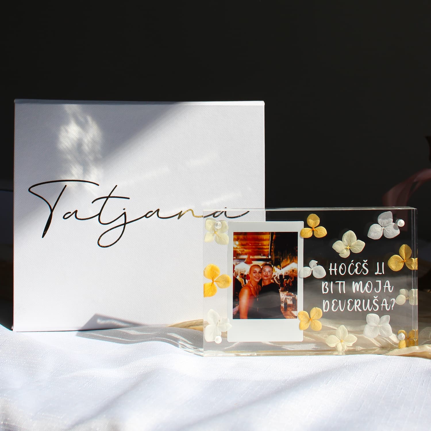 A clear acrylic photo frame with white and yellow flower decorations sits on a white fabric surface next to a white box with the name 'Tatjana' in black script. The photo inside the frame shows two people holding glasses, and the acrylic has the text 'HOCÍS LI BITI MOJA DEVRUSA?' in white lettering.