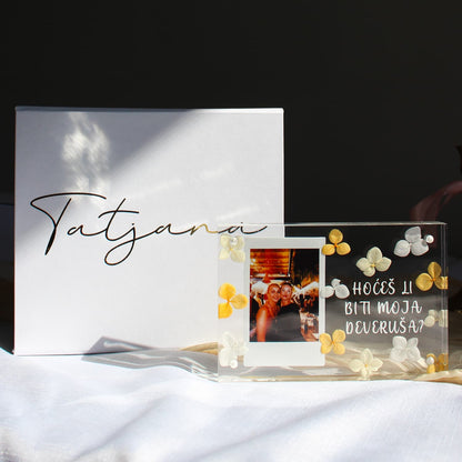 A clear acrylic photo frame with white and yellow flower decorations sits on a white fabric surface next to a white box with the name 'Tatjana' in black script. The photo inside the frame shows two people holding glasses, and the acrylic has the text 'HOCÍS LI BITI MOJA DEVRUSA?' in white lettering.