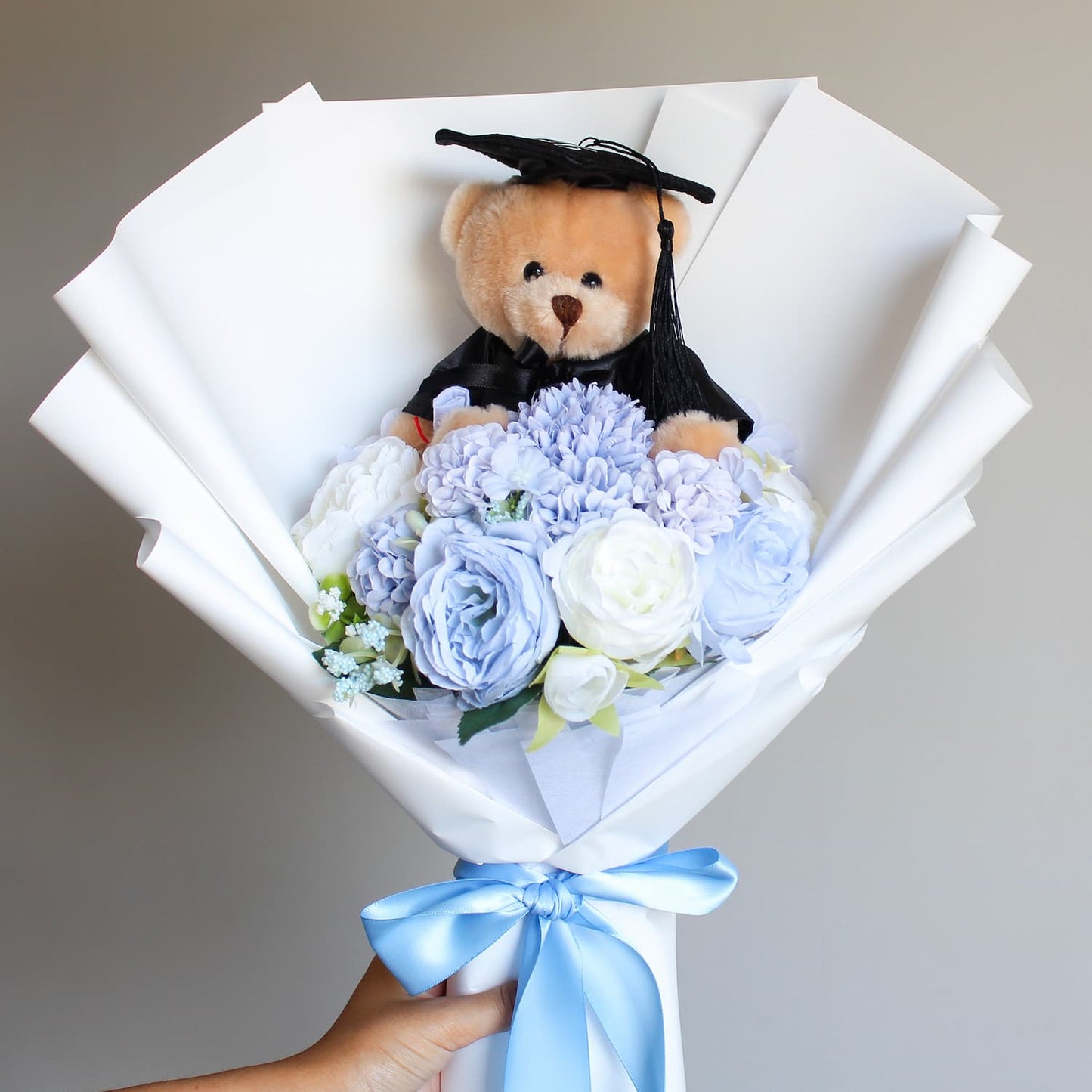 Graduation-themed teddy bear in a bouquet of flowers with a blue ribbon on a neutral background