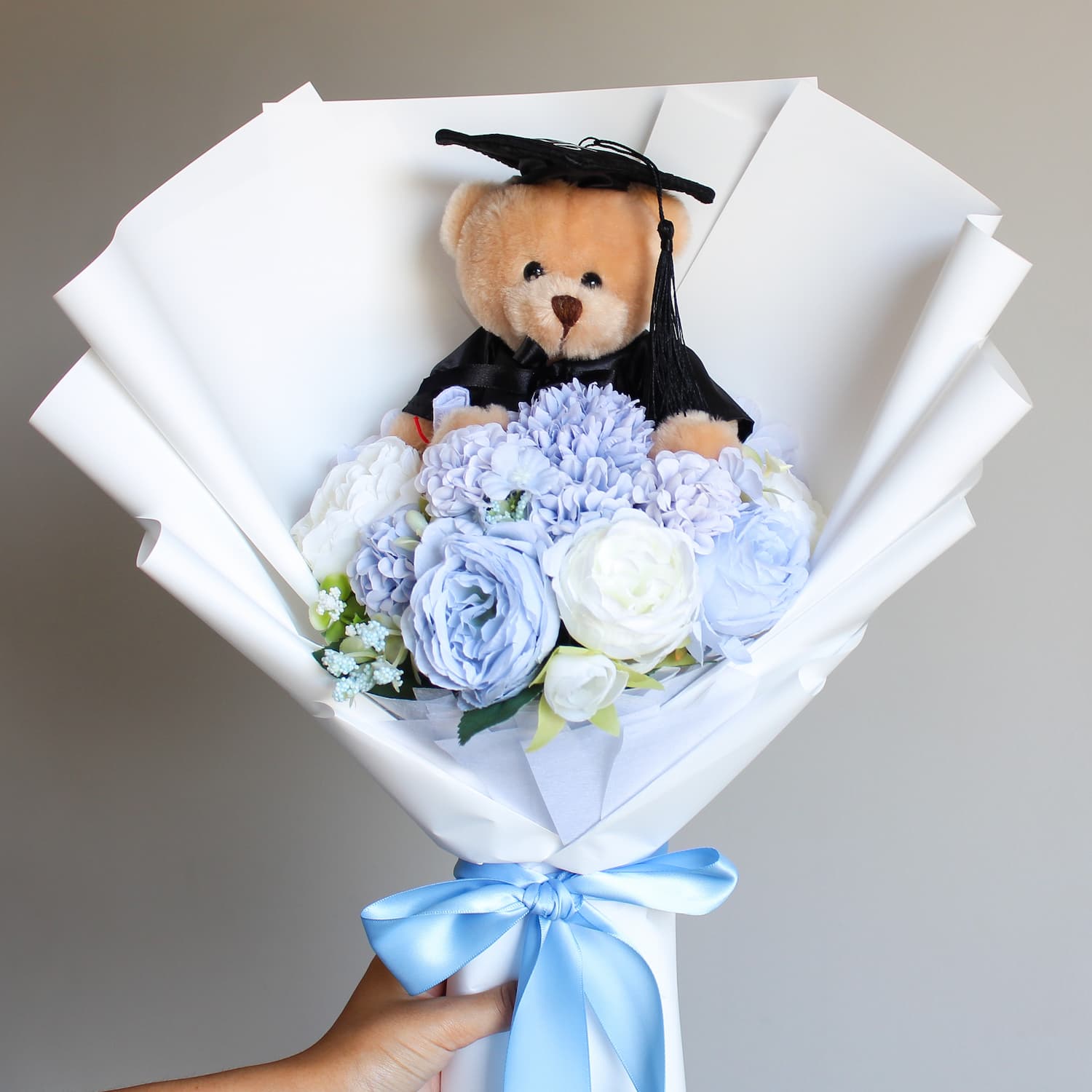 Graduation-themed teddy bear in a bouquet of flowers with a blue ribbon on a neutral background