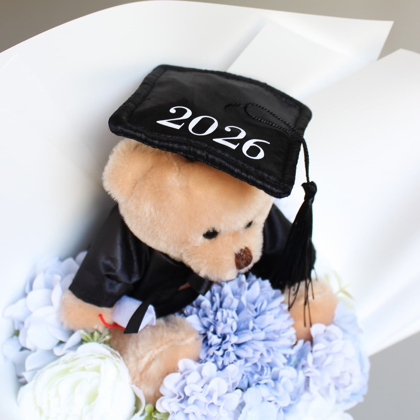 Teddy bear wearing a graduation cap with '2026' surrounded by flowers on a white background