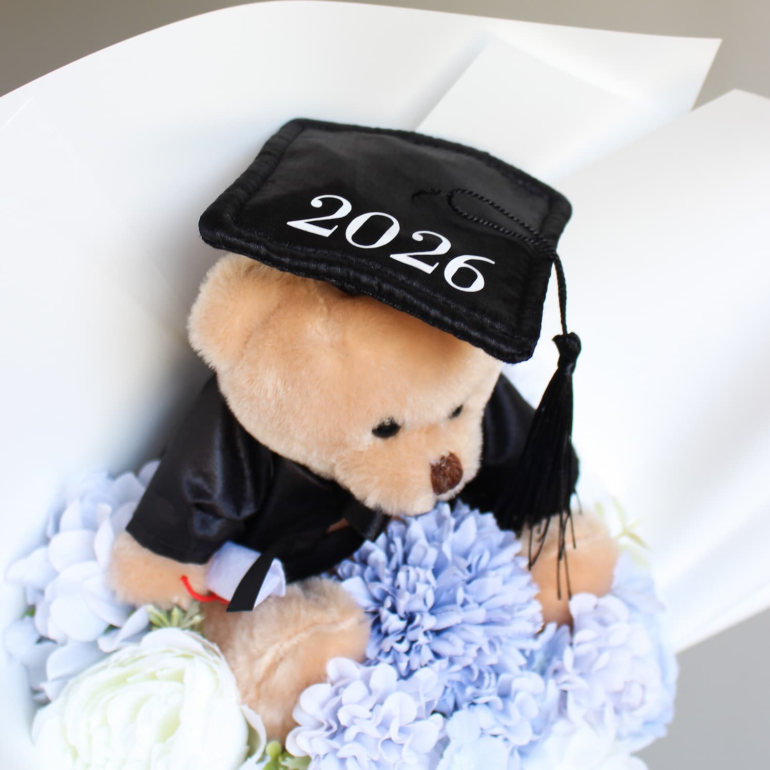 Teddy bear wearing a graduation cap with '2026' surrounded by flowers on a white background