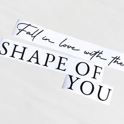 Two white rectangular decals with black text reading 'Fall in love with the SHAPE OF YOU' on a light gray background. personalised mirror decal
