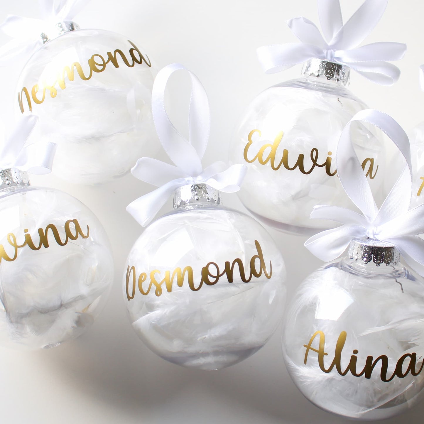 White feather Christmas ornaments with gold names and white ribbons on a light gray background