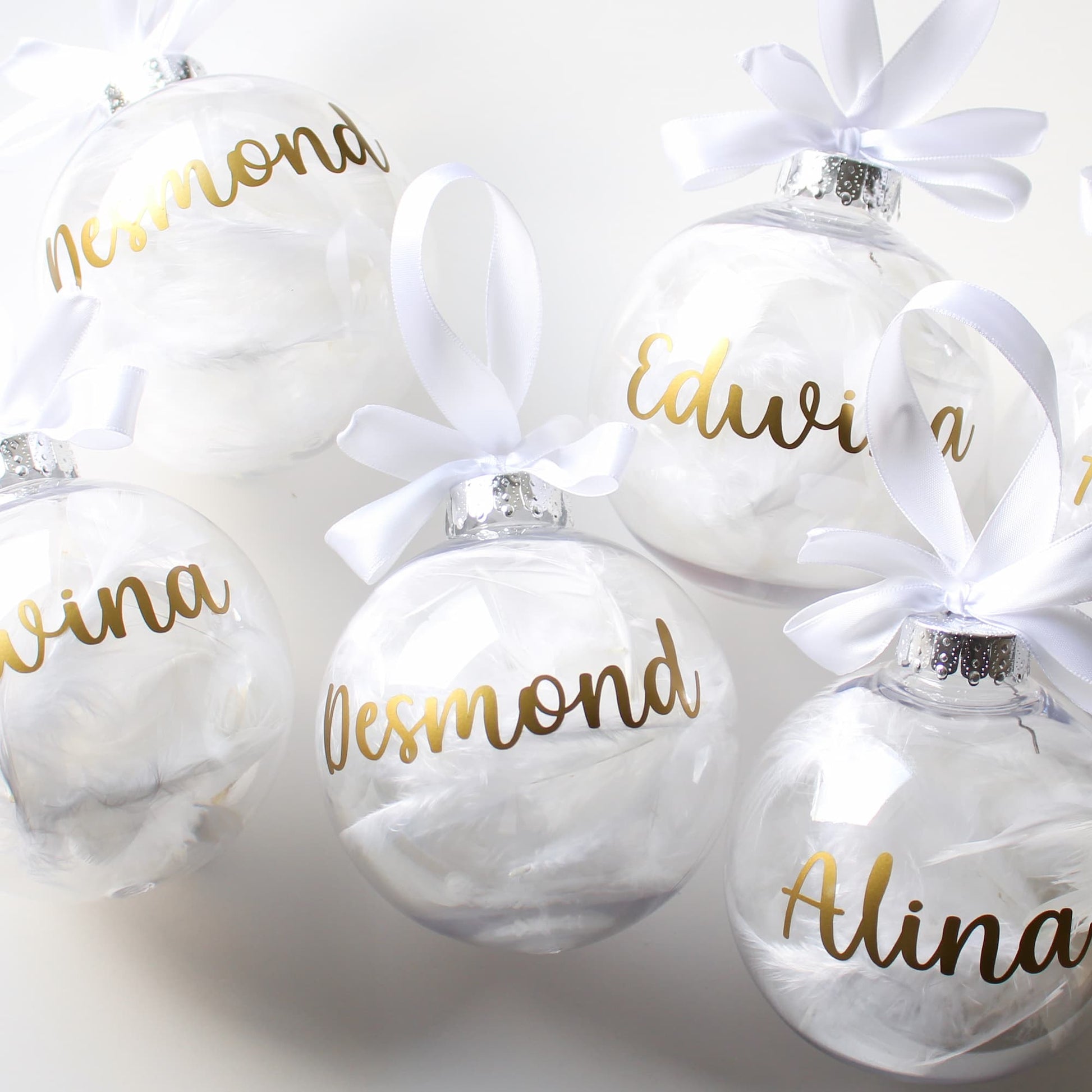 White feather Christmas ornaments with gold names and white ribbons on a light gray background