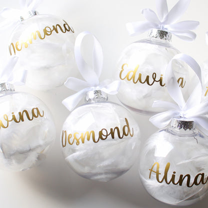 White feather Christmas ornaments with gold names and white ribbons on a light gray background