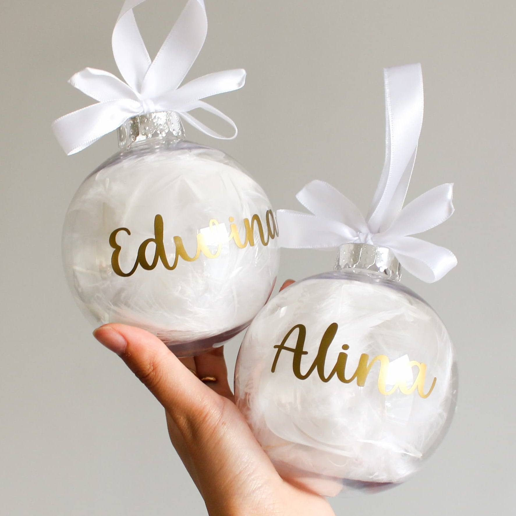 Two feather Christmas ornaments with gold names and white ribbons held by a hand against a neutral background.