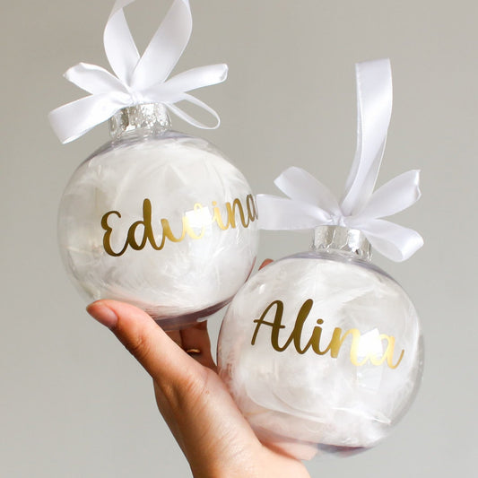 Two feather Christmas ornaments with gold names and white ribbons held by a hand against a neutral background.