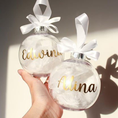 Two feather ornaments with white ribbons and gold names 'Edwin' and 'Alina' held by a hand.