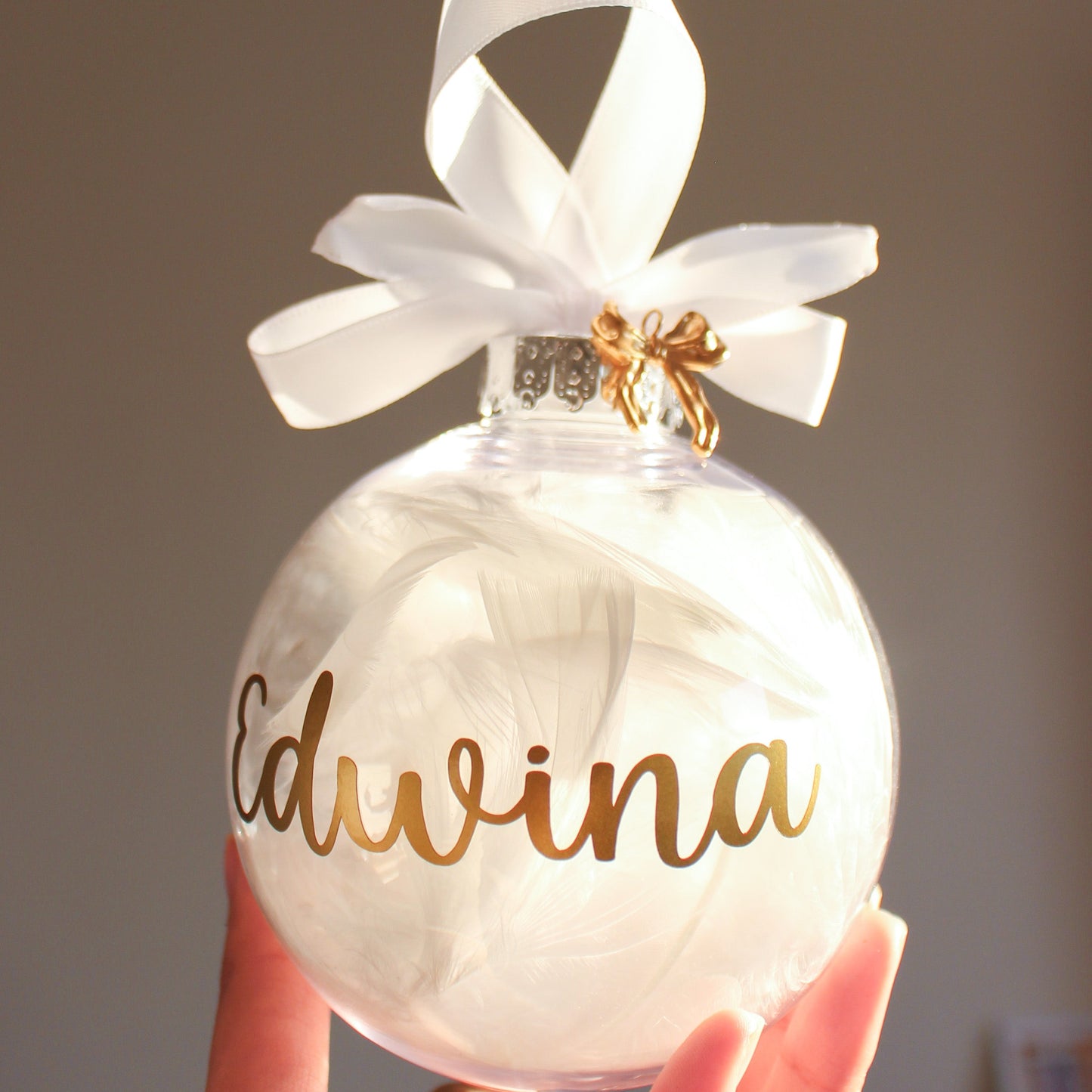 feather ornament with gold text 'Edwina' and a white ribbon, held by a hand against a neutral background.