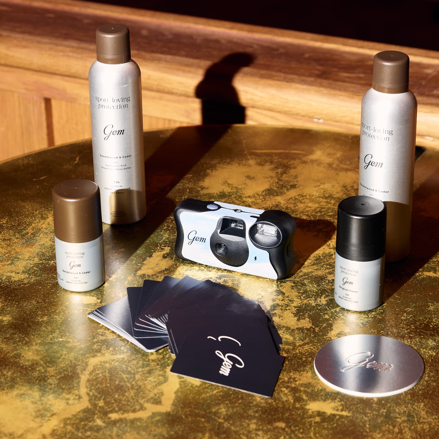 A set of Gem care products arranged on a gold-toned tabletop, including two tall bottles, two smaller bottles, a compact camera, cards, and a round tin, with a wooden surface in the background.