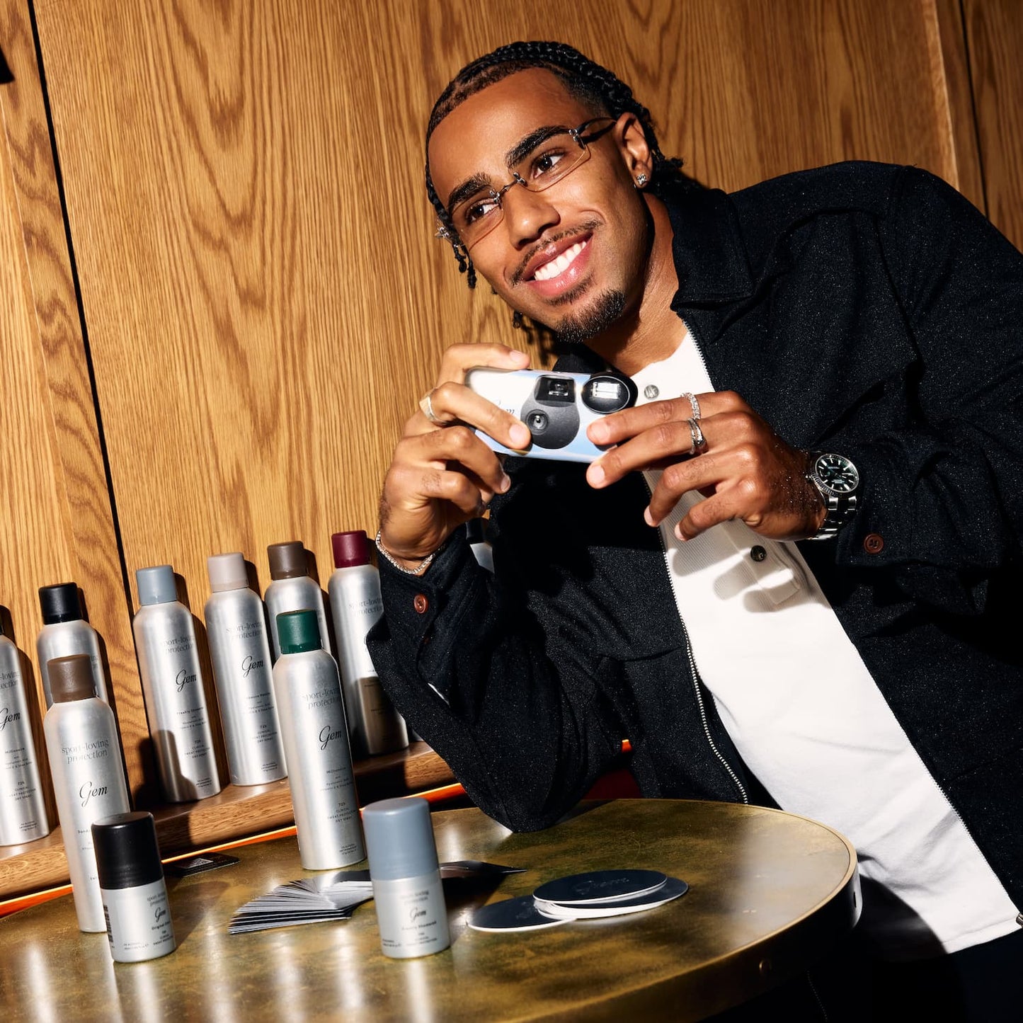 A man in a black jacket and white shirt holds a small camera in a warm, wood-paneled room, with multiple gem spray cans arranged on a table in the foreground.