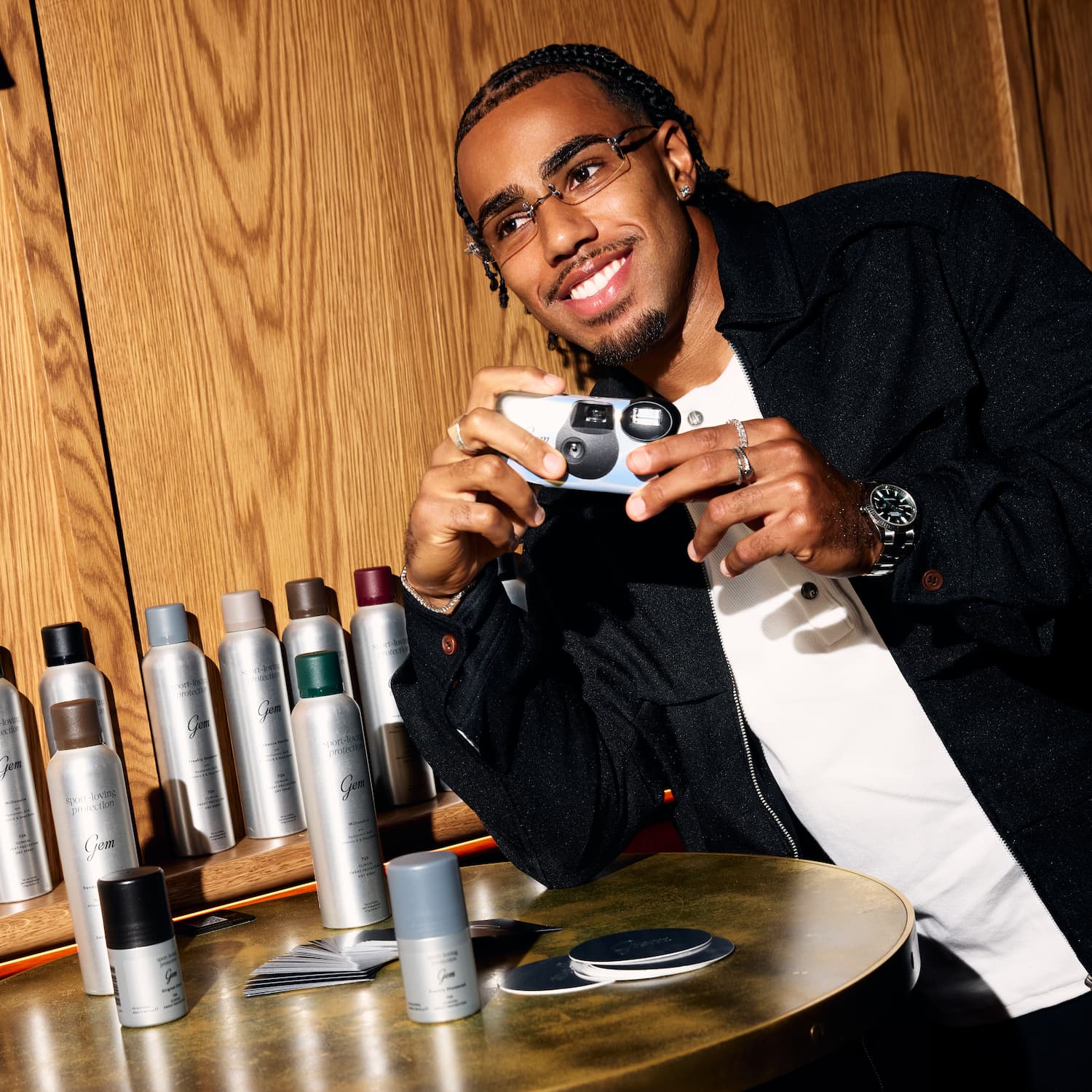 A man in a black jacket and white shirt holds a small camera in a warm, wood-paneled room, with multiple gem spray cans arranged on a table in the foreground.