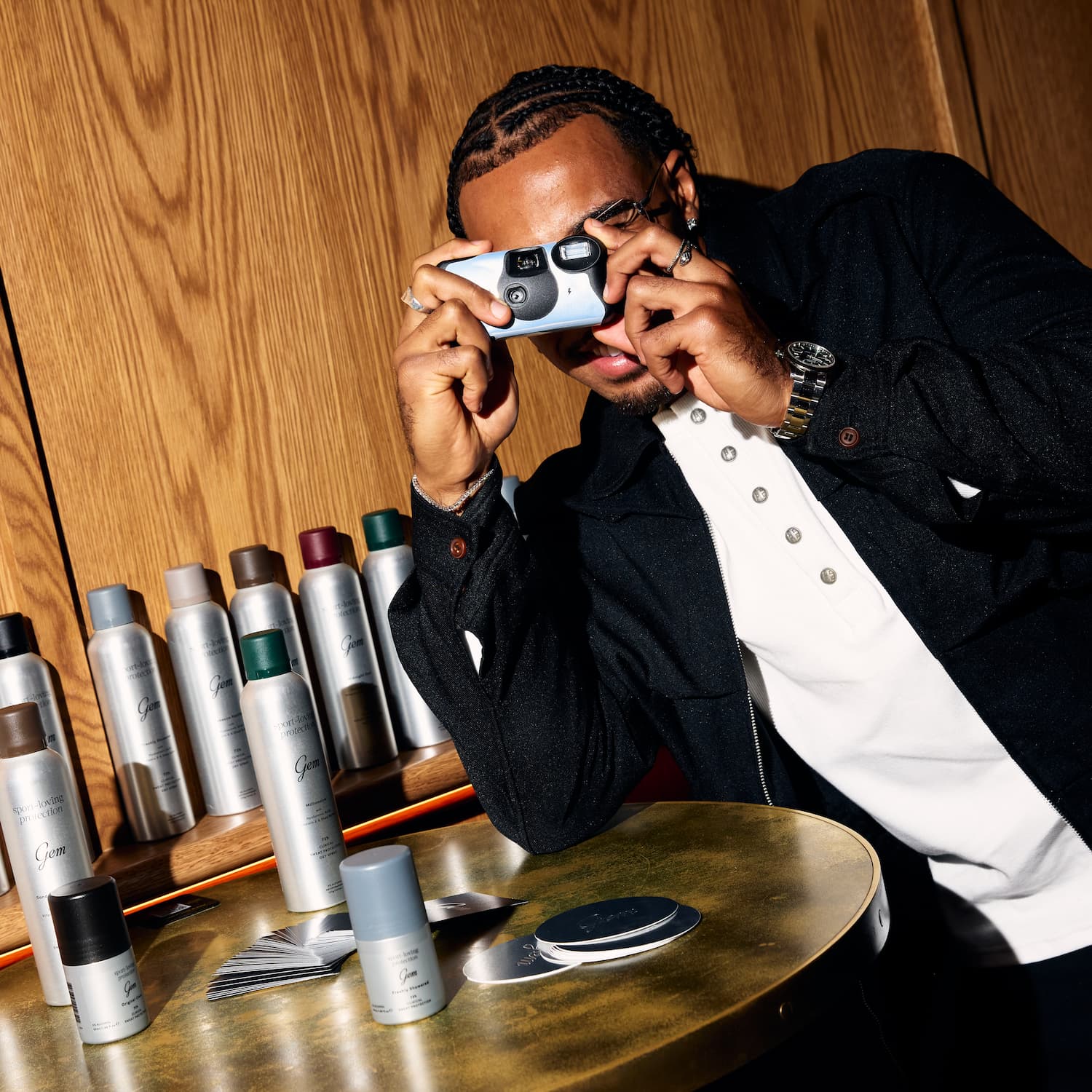 A man in a black jacket and white shirt holds a light blue camera up to his eyes, standing behind a gold table displaying various aerosol hairspray cans and hair products. The background is a warm wood-paneled wall.